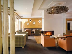 Lehrlingsportal.at Moderne Hotellounge mit Sesseln, Sofas, einem beleuchteten Kamin, Holzdekor, großen Fenstern und runden Spiegeln an einer Akzentwand aus Holz.