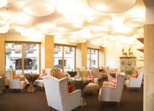 Lehrlingsportal.at Eine moderne Lounge mit beigen Sesseln, runden Glastischen, rot karierten Kissen, großen Fenstern, dekorativen Deckenleuchten und hölzernen Deko-Akzenten.