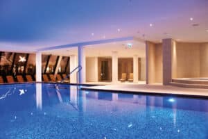 Lehrlingsportal.at Hallenbadbereich mit blau gefliestem Becken, Liegestühlen und stimmungsvoller Beleuchtung, mit Säulen und einem Sitzbereich im Hintergrund.