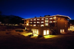 Lehrlingsportal.at Ein großes, modernes Gebäude mit beleuchteten Fenstern und Lichterketten ist bei Nacht vor einem dunkelblauen Himmel abgebildet, wobei die Berge im Hintergrund schwach zu erkennen sind.