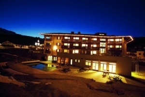 Lehrlingsportal.at Ein hell erleuchtetes Hotelgebäude bei Nacht, mit Balkonen, großen Fenstern und einem Außenpool, vor einem tiefblauen Himmel in einer Bergregion.