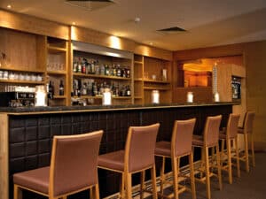 Lehrlingsportal.at Eine moderne Bar mit schwarzer, gepolsterter Front, Holzregalen mit Flaschen und Gläsern und sechs braunen Barhockern, die ordentlich aufgereiht sind.