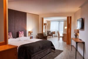 Lehrlingsportal.at Modernes Hotelzimmer mit Doppelbett, Sesseln, Schreibtisch, Lampen, Flachbildfernseher und großen Fenstern mit transparenten Vorhängen, in neutralen Farbtönen gehalten.