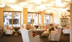 Lehrlingsportal.at Eine moderne Lounge mit cremefarbenen Sesseln, Glastischen mit Holzgestell, großen Fenstern, karierten Kissen und dekorativen Hirschstatuen in einem hellen, gemütlichen Ambiente.