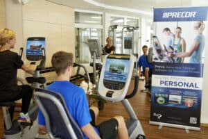Lehrlingsportal.at Vier Personen benutzen stationäre Fahrräder in einem Fitnessstudio neben einem großen Precor-Werbebanner für Fitnesssoftware, mit Trainingsgeräten im Hintergrund.