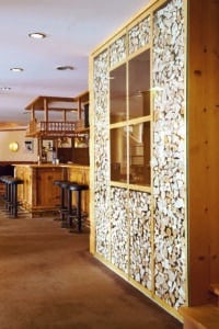 Lehrlingsportal.at Ein hölzerner Barbereich mit schwarzen Hockern steht neben einer dekorativen Wand mit gestapelten Holzstücken auf einem braunen Teppichboden in einem hell erleuchteten Raum.