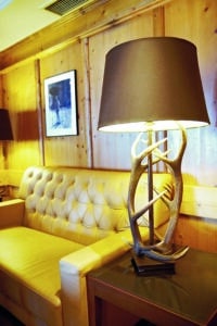 Lehrlingsportal.at Ein hellbraunes Ledersofa steht an einer holzgetäfelten Wand, darüber ein gerahmtes Foto; auf einem Beistelltisch steht eine Tischlampe mit einem geweihförmigen Sockel und einem dunklen Schirm.