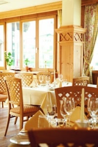 Lehrlingsportal.at Ein gut beleuchteter Restaurantbereich mit Holzstühlen, weiß gedeckten Tischen, Gläsern und gefalteten Servietten in der Nähe großer Fenster mit Vorhängen.