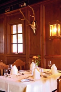 Lehrlingsportal.at Ein ordentlich gedeckter Esstisch mit gefalteten Servietten und Gläsern in einem holzgetäfelten Restaurant, dekoriert mit einem an der Wand befestigten Geweihschädel und einer Laterne im Stil einer Lampe.