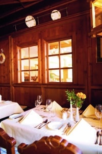 Lehrlingsportal.at Ein ordentlich gedeckter Restauranttisch mit weißem Tischtuch, Servietten, Gläsern, Besteck und einem kleinen Blumenarrangement, daneben eine Holzwand mit Fenstern und dekorativen Platten.