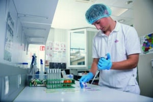 Lehrlingsportal.at Eine Person in einem Laborkittel und mit Haarnetz benutzt eine Pipette an einem Laborarbeitsplatz mit Reagenzgläsern und Laborgeräten.