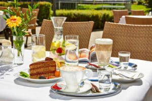 Lehrlingsportal.at Ein Cafétisch im Freien, gedeckt mit einem Stück Kuchen, Cappuccino, Milchkaffee, Wassergläsern, einer Karaffe mit Fruchtwasser und einer Blumenvase an einem sonnigen Tag.