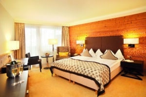 Lehrlingsportal.at Modernes Hotelzimmer mit einem großen Bett, einer Akzentwand aus Holz, einem Sessel, einem kleinen Tisch, einem Schreibtisch und einem Fenster mit Vorhängen, die natürliches Licht hereinlassen.