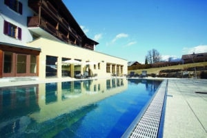 Lehrlingsportal.at Außenschwimmbad neben einem mehrstöckigen Gebäude mit Balkonen, Liegestühlen und fernen Bergen unter einem klaren blauen Himmel.