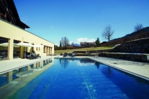 Lehrlingsportal.at Außenschwimmbad neben einem Gebäude, mit Liegestühlen, Sonnenschirmen und einer Berglandschaft im Hintergrund unter blauem Himmel.