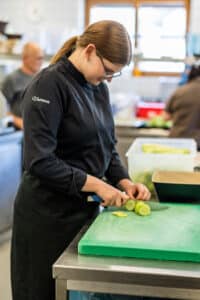 Lehrlingsportal.at Ein Koch in schwarzer Uniform mit der Aufschrift "Chelsea" schneidet Gurken auf einem grünen Schneidebrett in einer Großküche.