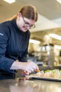 Lehrlingsportal.at Ein Koch in schwarzer Uniform und mit Gläsern garniert sorgfältig Teller mit Speisen in einer professionellen Küche.