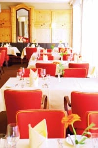 Lehrlingsportal.at Ein heller Restaurantbereich mit roten Stühlen, weißen Tischtüchern, ordentlich gefalteten Servietten und Gläsern auf Tischen in der Nähe großer Fenster.