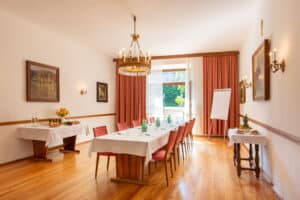 Lehrlingsportal.at Ein heller Tagungsraum mit einem langen Tisch, Stühlen, einem Flipchart und Tischen mit Snacks und Getränken, dekoriert mit Bildern und einem Kronleuchter.