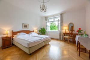 Lehrlingsportal.at Ein ordentliches Hotelzimmer mit zwei Betten, Holzmöbeln, einem Kronleuchter, einem Tisch mit Blumen und einem offenen Fenster, durch das man ins Grüne schauen kann.