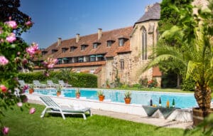 Lehrlingsportal.at Außenschwimmbad mit Liegestühlen, Topfpflanzen und Getränken in einem Garten neben einem historischen Steingebäude mit Ziegeldach und Bogenfenstern.