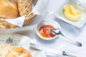 Lehrlingsportal.at Ein Frühstückstisch mit einem Brotkorb, einer Schale mit Butter, einer Schale mit Marmelade und einem Löffel sowie einem Brötchen auf einem Teller.