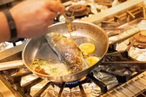 Lehrlingsportal.at Eine Person kocht einen ganzen Fisch in einer Pfanne mit Zitronenscheiben und Kräutern auf einem Gasherd und begießt ihn mit einem Löffel.
