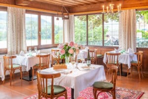 Lehrlingsportal.at Ein heller Restaurantbereich mit Holzstühlen, weißen Tischtüchern, Blumenschmuck, großen Fenstern und Bergblick im Hintergrund.