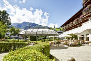 Lehrlingsportal.at Hotel-Essbereich im Freien mit Tischen und Stühlen unter Sonnenschirmen und einem Baldachin, umgeben von Grün und Blumen, mit Bergen und blauem Himmel im Hintergrund.