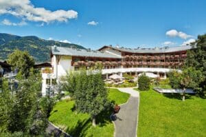 Lehrlingsportal.at Ein großes Hotel mit Balkonen und Blumenkästen überblickt eine grüne Wiese und einen Garten vor der Kulisse der Berge und des klaren blauen Himmels.