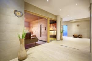 Lehrlingsportal.at Modernes Spa-Interieur mit hellen Fliesen, Saunen mit Glastüren, Topfpflanzen, hölzernen Dekorationsstücken und versenkten Deckenleuchten.