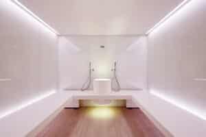 Lehrlingsportal.at Minimalistisches Dampfbad mit weißen Bänken, zwei Handbrausen an gegenüberliegenden Wänden, einer runden weißen Mittelarmatur und sanfter Umgebungsbeleuchtung.