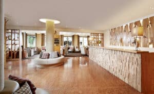 Lehrlingsportal.at Geräumige Hotellobby mit holzgetäfelter Rezeption, runder Sitzecke, dekorativen Kissen und großen Fenstern, die für Tageslicht sorgen.
