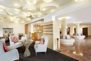 Lehrlingsportal.at Moderner Loungebereich mit Sesseln, rundem Tisch und karierten Kissen, mit Deckenleuchten, Holzregalen und einem Empfangstresen im Hintergrund.