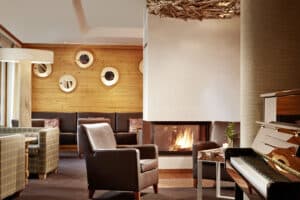 Lehrlingsportal.at Gemütliche Hotellounge mit Ledersesseln, einem Klavier, einem modernen Kamin, holzgetäfelten Wänden und runden Spiegeln als Dekoration.