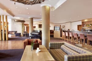 Lehrlingsportal.at Moderne Hotellobby mit kariertem Sofa, Holztisch, Kerze, Klavier, Kamin und einem Barbereich mit Hochstühlen im Hintergrund.