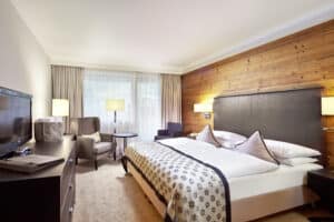 Lehrlingsportal.at Modernes Hotelzimmer mit einem großen Bett, gemusterter Bettwäsche, zwei Sesseln, einem Flachbildfernseher, einer Akzentwand aus Holz und einem großen Fenster mit Vorhängen.