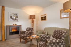 Lehrlingsportal.at Ein gemütliches Wohnzimmer mit einem beleuchteten Kamin, zwei gemusterten Sesseln, einem grünen Sofa, einem hölzernen Couchtisch mit Büchern und Wandkunst an weißen Wänden.