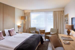 Lehrlingsportal.at Ein modernes Hotelzimmer mit einem Doppelbett, zwei Sesseln am Fenster, einem Holzschreibtisch mit Stuhl, sanfter Beleuchtung und neutralen Farbtönen.