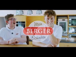 Lehrlingsportal.at Zwei junge Männer in weißen Hemden sitzen an einem Küchentisch; der eine lächelt und lacht, der andere schaut nachdenklich. Der Schriftzug "Mein BERGER Schinken" ist über das Bild gelegt.