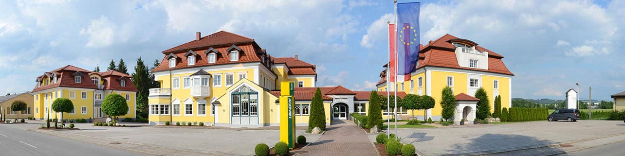 Lehrlingsportal.at Panoramablick auf einen gelben Gebäudekomplex mit roten Dächern, Flaggen der Europäischen Union, Grünflächen und einer gepflasterten Auffahrt unter einem teilweise bewölkten Himmel.