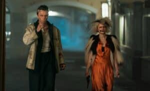 Lehrlingsportal.at Zwei Personen in Halloweenkostümen, eine als Frankensteins Monster in zerschlissenen Kleidern und die andere in einem orangefarbenen Kleid mit dramatischem Make-up, gehen eine schwach beleuchtete Straße entlang.