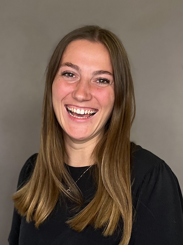 Lehrlingsportal.at Eine Frau mit langem glattem braunem Haar und schwarzem Oberteil lächelt breit vor einem schlichten grauen Hintergrund.