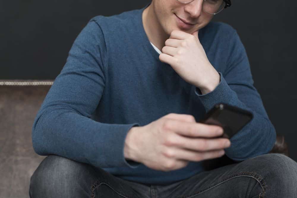 Lehrlingsportal.at Eine Person mit blauem Pullover und Brille sitzt im Schneidersitz und schaut mit einem leichten Lächeln auf ein Smartphone.