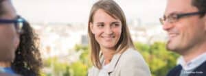Lehrlingsportal.at Drei Personen befinden sich im Freien, wobei eine Frau in der Mitte lächelt und einen Mann auf der rechten Seite anschaut. Der Hintergrund ist unscharf mit Grünflächen und Gebäuden.