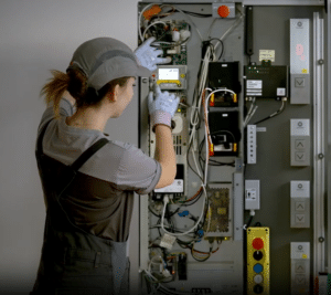 Lehrlingsportal.at Ein Techniker in Uniform und Handschuhen justiert Komponenten in einer offenen elektrischen Schalttafel mit verschiedenen Kabeln, Schaltern und Geräten.