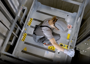 Lehrlingsportal.at Ein Arbeiter in grauem Overall und Handschuhen kniet in einem Industrieaufzugsschacht und bedient die Bedienelemente auf einer Metallplattform mit Warnschildern und Sicherheitsetiketten.