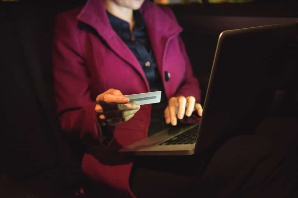 Lehrlingsportal.at Person in einem kastanienbraunen Mantel, die eine Kreditkarte in der Hand hält und einen Laptop benutzt, in einem Innenraum bei schwacher Beleuchtung sitzend.