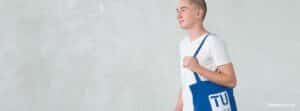 Lehrlingsportal.at Eine Person in einem weißen T-Shirt hält eine blaue Tragetasche mit dem Aufdruck "TU" in der Hand und steht vor einem schlichten hellen Hintergrund.