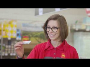 Lehrlingsportal.at Eine Frau in roter Uniform und mit Brille lächelt, während sie in einem Geschäft eine Karte hochhält. Im Hintergrund sind Regale mit Produkten zu sehen.